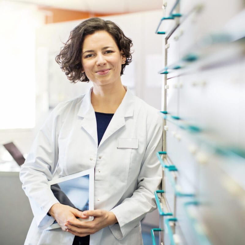 Portrait of confident young female provider leaning to a medicine shelf with a digital tablet