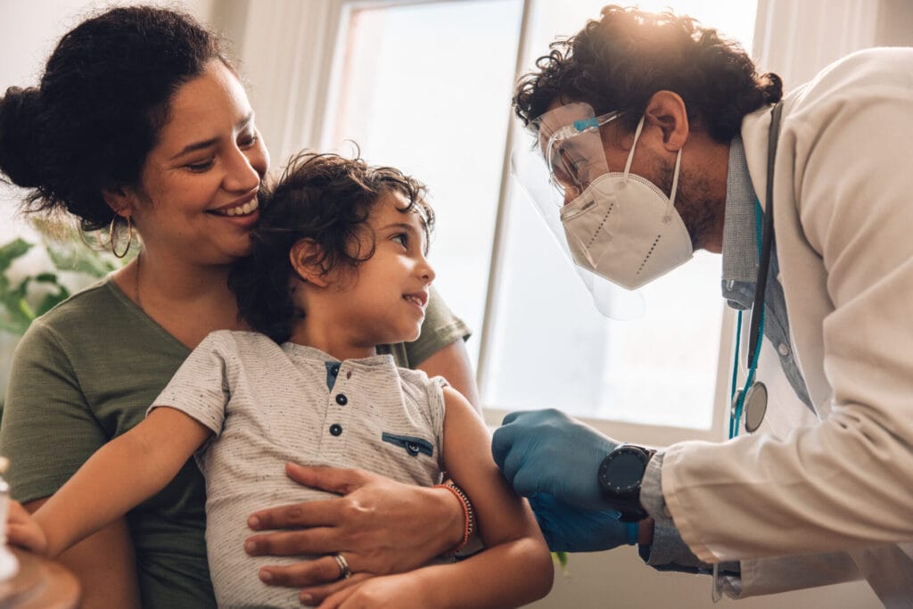 Doctor putting a bandage on arm of a boy after giving vaccine. Boy looking at frontline worker and smiling after getting vaccination.